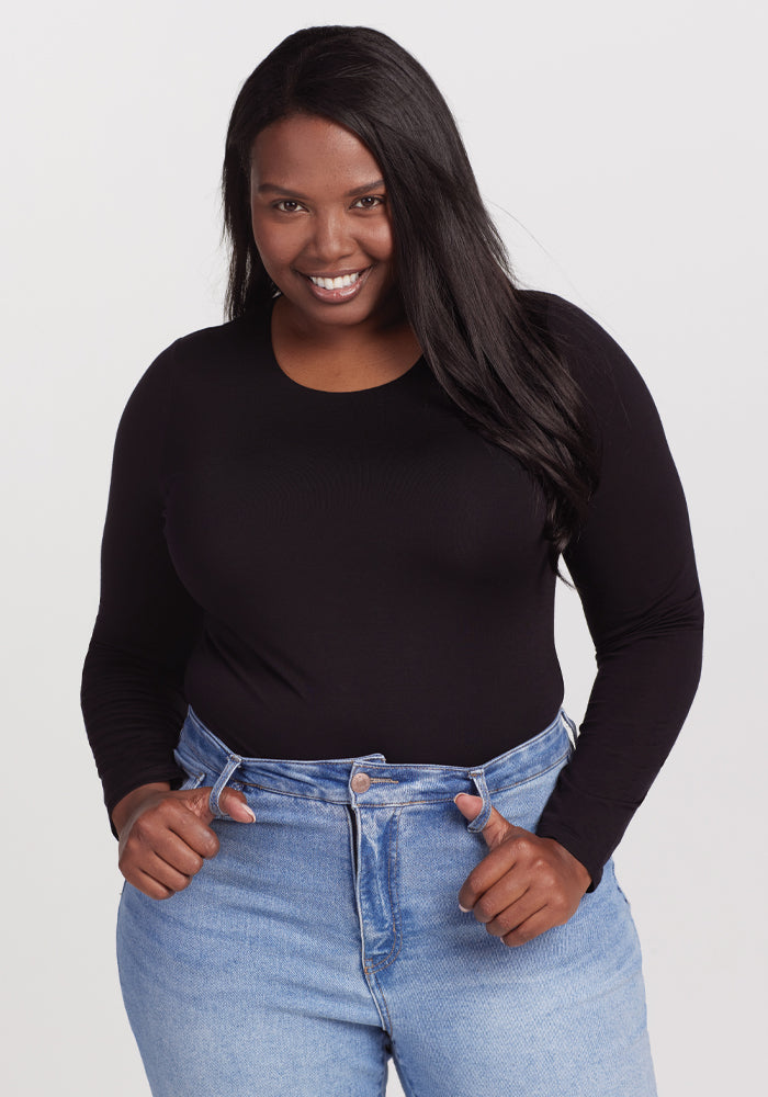 A person smiling, dressed in a Woolx Sophia Bodysuit - Black and high-waisted blue jeans. Their long, dark hair flows naturally as they stand against a simple white background. | Le'Quita is 5'11", wearing a size XL