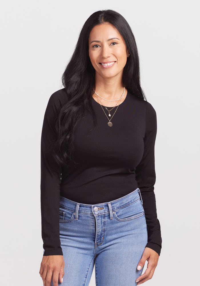 A person with long black hair and a relaxed demeanor wears a Woolx Sophia Bodysuit in black and light blue jeans, smiling against a plain white background. They've layered necklaces for style. | Denia is 5'8", wearing a size S