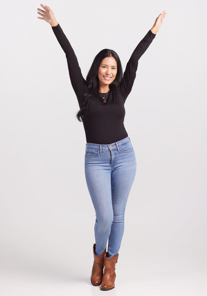 A woman with long black hair smiles and raises her arms upward, wearing the Woolx Sophia Bodysuit in black, light blue jeans, and brown ankle boots. She stands against a plain white background.