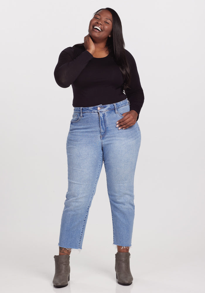 A person in a Woolx Sophia Bodysuit - Black, light blue high-waisted jeans, and gray ankle boots smiles with a hand on their neck against a plain white background.