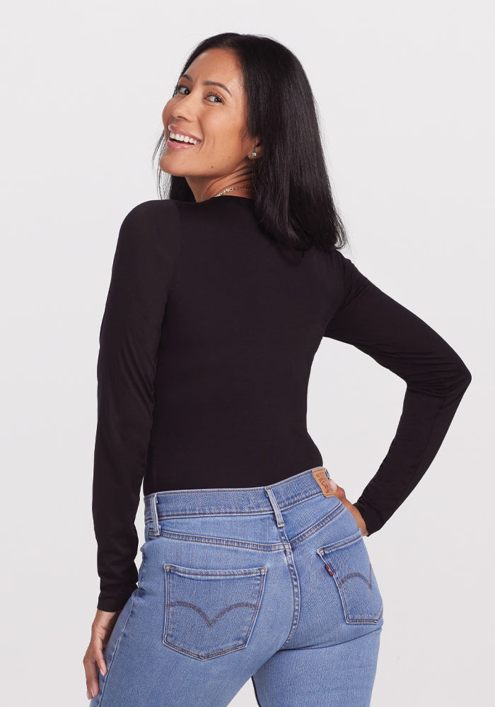 A smiling woman with long black hair wears the Woolx Sophia Bodysuit in black and blue jeans, standing with her back to the camera, looking over her shoulder against a white background.