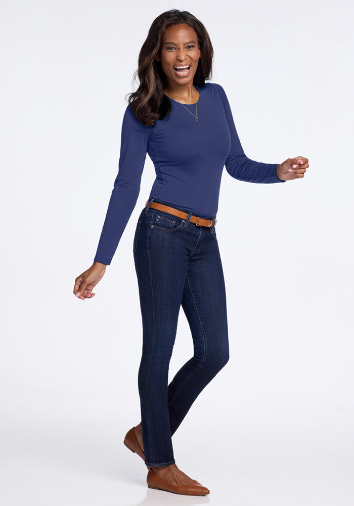A playful individual poses against a light background, wearing the Woolx Sophia Bodysuit in Blue Horizon with dark jeans and brown shoes. Their hair is loose, styled with a necklace and brown belt, exuding a casual and cheerful vibe.