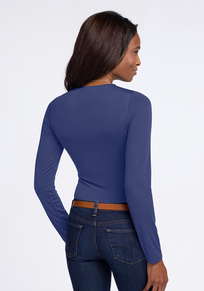 A person with long hair stands facing away, dressed in the Sophia Bodysuit - Blue Horizon by Woolx and dark jeans. The plain white background accentuates the outfit's stylish simplicity.