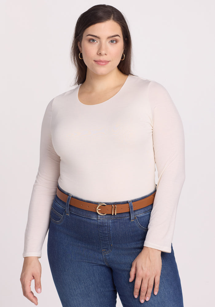 A woman with long dark hair models a chic outfit against a plain backdrop, featuring the Sophia Bodysuit in Vanilla by Woolx paired with blue jeans and a brown belt. | Juliana is 5'10", wearing a size XL