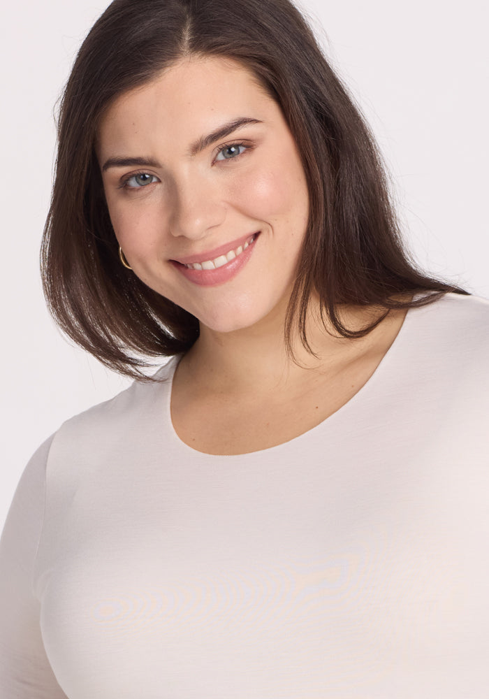 A smiling woman with long dark hair wears the Vanilla Sophia Bodysuit by Woolx, looking directly at the camera against a plain white background.