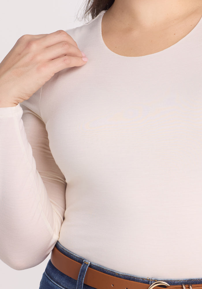 A person wearing a fitted long-sleeve Woolx Sophia Bodysuit - Vanilla adjusts the neckline while also sporting blue jeans and a brown belt. The focus is on the torso and hand, with no face visible.