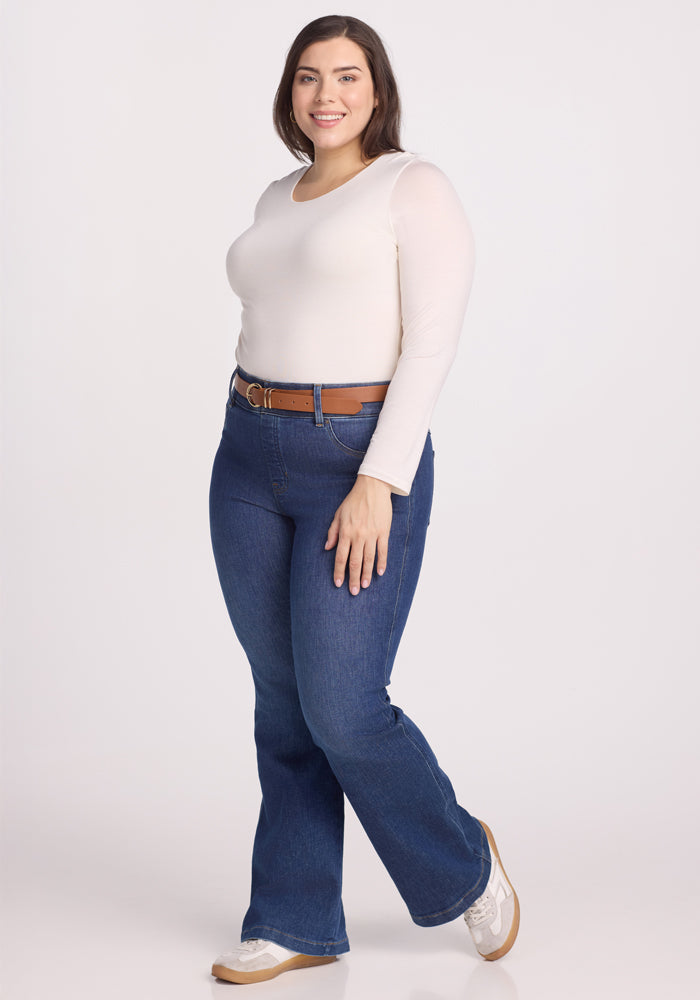 A person with shoulder-length brown hair smiles while standing in a Woolx Sophia Bodysuit - Vanilla, blue jeans, beige sneakers, and a brown belt against a plain white background.