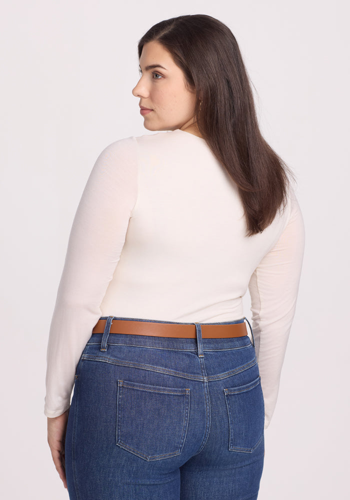 A person with long, dark hair is seen from the back, wearing the Woolx Sophia Bodysuit in Vanilla and blue jeans with a brown belt. They stand gracefully against a plain white background.