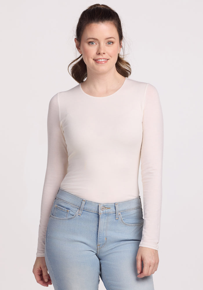 A person with long hair wearing a Woolx Sophia Bodysuit in Vanilla and light blue jeans stands smiling gently at the camera against a plain background. | Lilly is 5’9”, wearing a size S