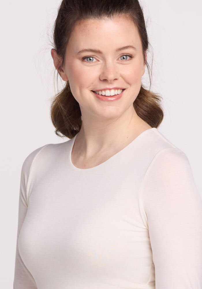 A smiling person with medium-length brown hair wears a vanilla Sophia Bodysuit by Woolx. They face the camera against a neutral background, radiating effortless style.