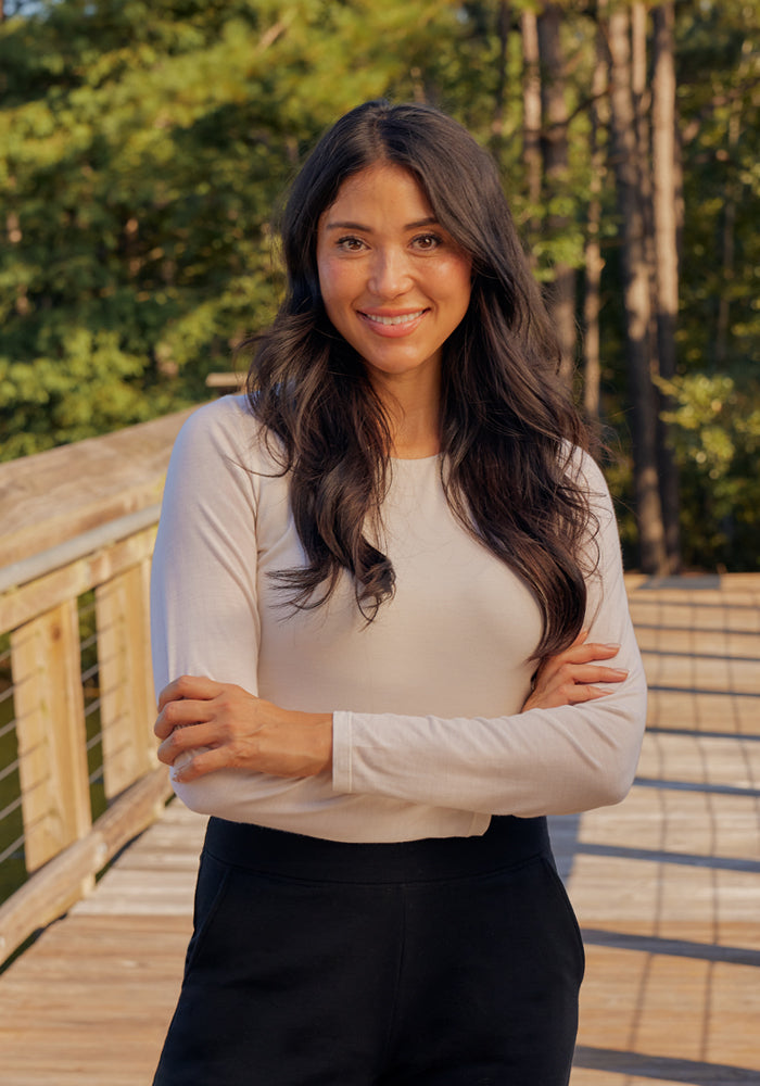 A woman with long dark hair wears a white Woolx Sophia Bodysuit and black pants, standing on a wooden bridge outdoors with her arms crossed and smiling, surrounded by green trees.