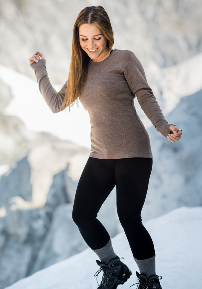A woman with long brown hair, wearing a brown long-sleeve shirt, Woolx Stella Leggings in black, gray socks, and black boots, smiles and balances on snow with a blurred mountain background.