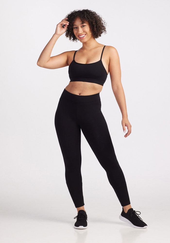 A person in Stella Leggings - Black by Woolx and a black sports bra with a sleek contouring fit stands smiling, one hand touching their curly hair. They are wearing black athletic shoes and posing against a plain white background.
