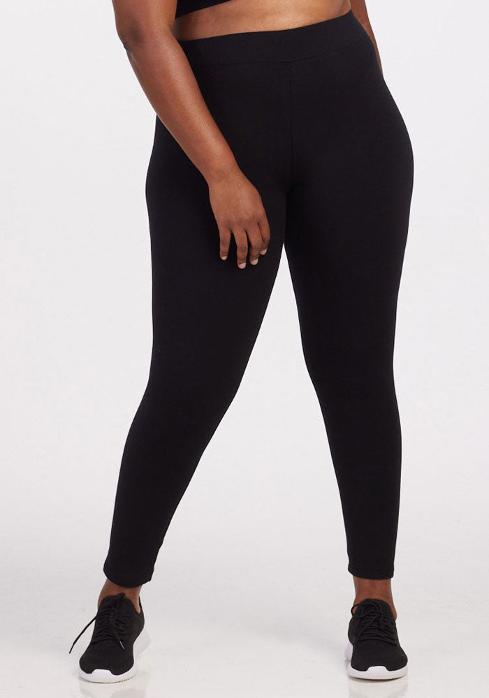 A person wearing the Woolx Stella Leggings - Black, featuring flattering back seams, along with black sneakers stands against a plain background, their left hand resting casually on their thigh. | Le'Quita is 5'11", wearing a size XL