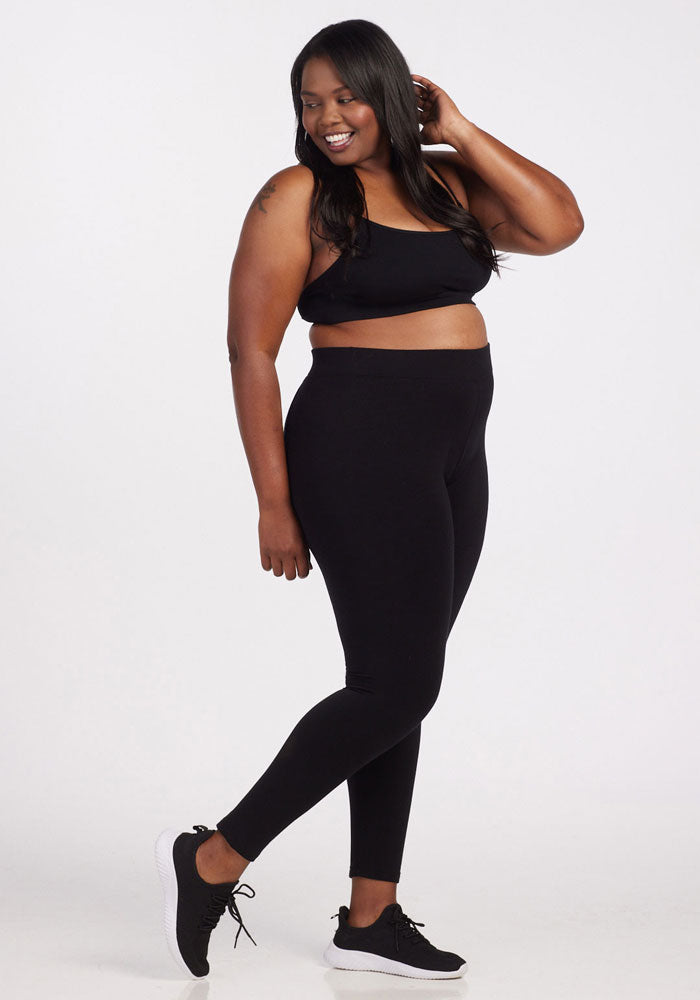 Dressed in Woolx's Stella Leggings - Black, paired with a black sports bra and sneakers, a person is posing and smiling. The flattering back seams and sleek contouring fit of the leggings enhance their appearance against the minimalist white studio background.