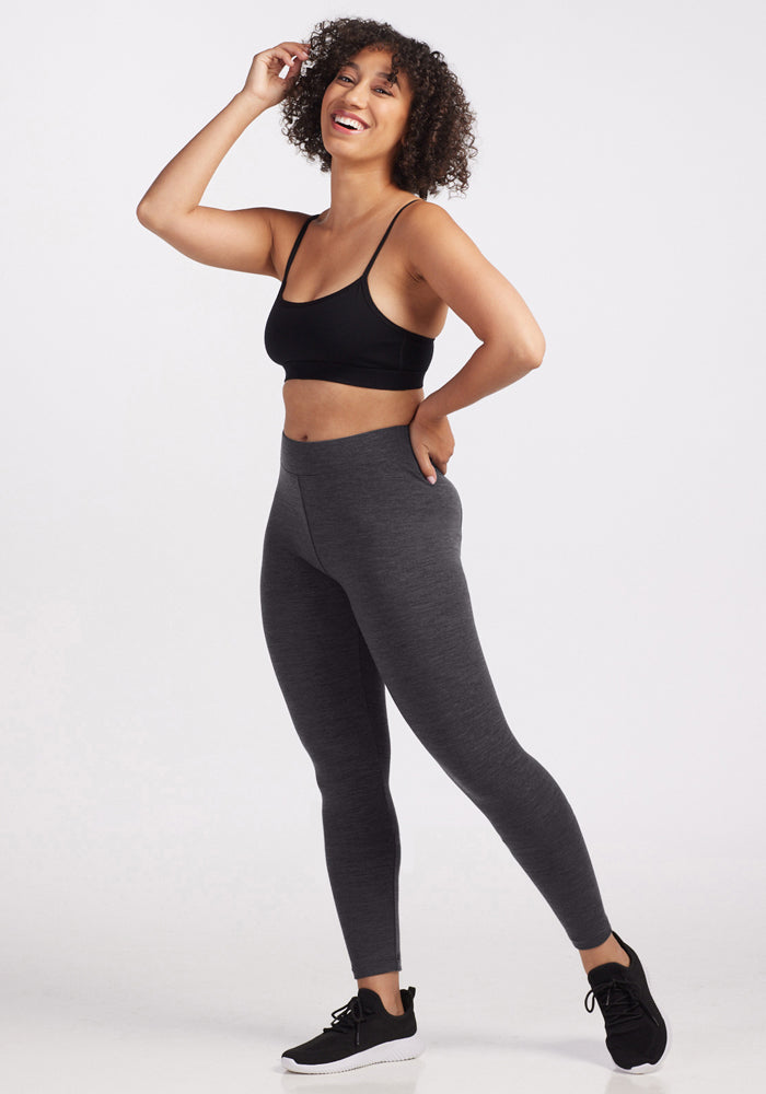 A woman with curly hair smiles while wearing Woolx Stella Leggings in Cinder Melange, a black sports bra, and black sneakers. She stands against a plain white background, one hand in her pocket and the other touching her hair.