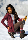 A woman smiles with eyes closed, kneeling in the snow in Woolx Stella Leggings • Final Sale - Bracken, a maroon hoodie, hiking boots, colorful socks, and holding tan gloves. Snowy trees blur softly in the background.