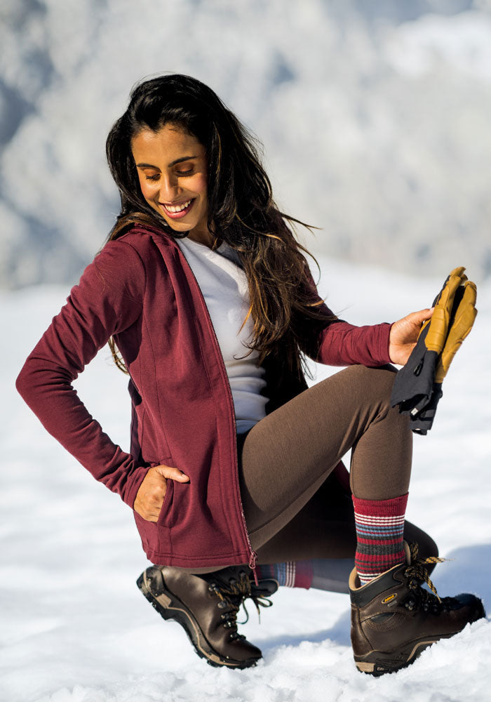 A woman smiles with eyes closed, kneeling in the snow in Woolx Stella Leggings • Final Sale - Bracken, a maroon hoodie, hiking boots, colorful socks, and holding tan gloves. Snowy trees blur softly in the background.