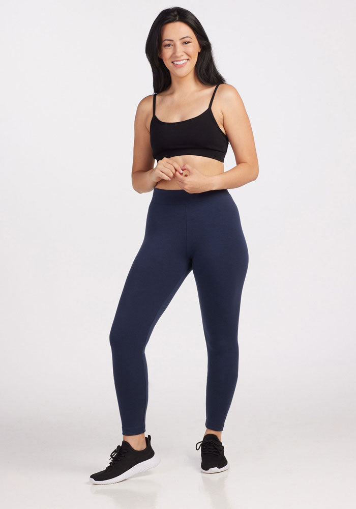A woman with long dark hair stands smiling against a plain background, wearing Woolx Stella Leggings in Deep Navy (Final Sale) and a black sports bra, with her hands clasped in front of her.