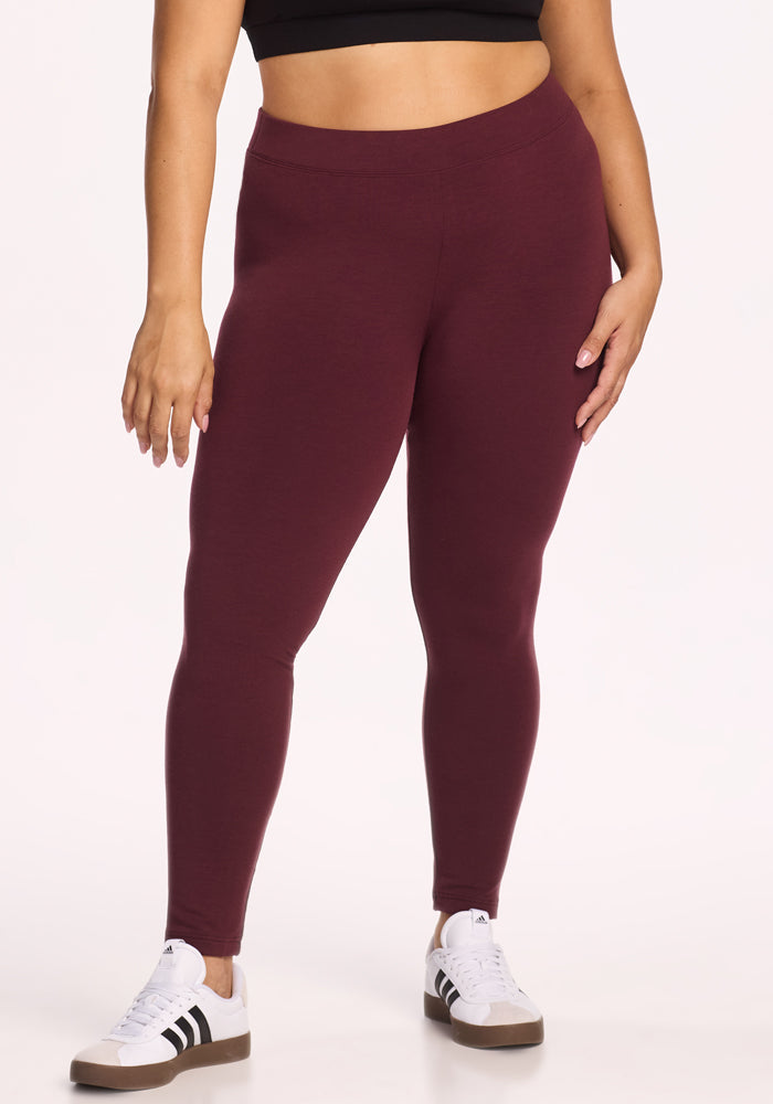 A person wearing Woolx Stella Leggings in Toasted Garnet, a black crop top, and white sneakers with black stripes stands against a plain light background.