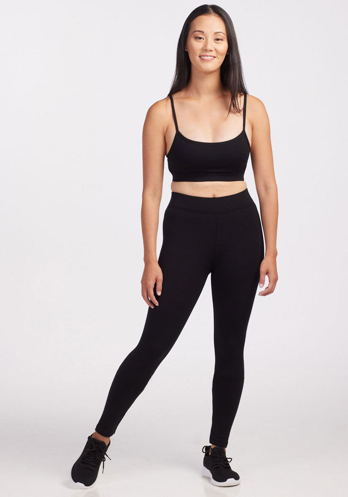 A person stands smiling, donned in the Stella Petite - Black sports bra and leggings by Woolx, crafted from DuraLite fabric, and paired with black sneakers. The backdrop is a plain white.