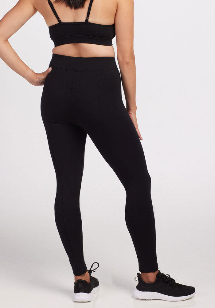A person dressed in a Stella Petite - Black sports bra and high-waisted leggings crafted from DuraLite fabric by Woolx, along with matching black sneakers, stands with their hand on their hip against a plain white background.