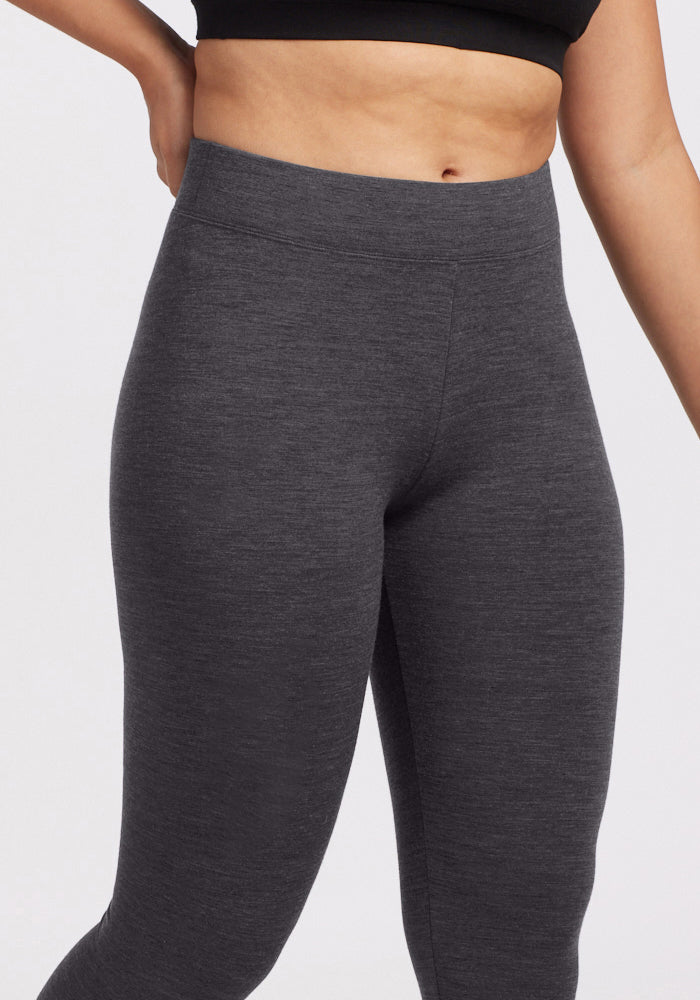 A person wears Woolx Stella Petite - Cinder Melange high-waisted dark gray merino wool leggings with a black crop top, standing with one hand on their hip, partially showing their torso and legs against a plain background.