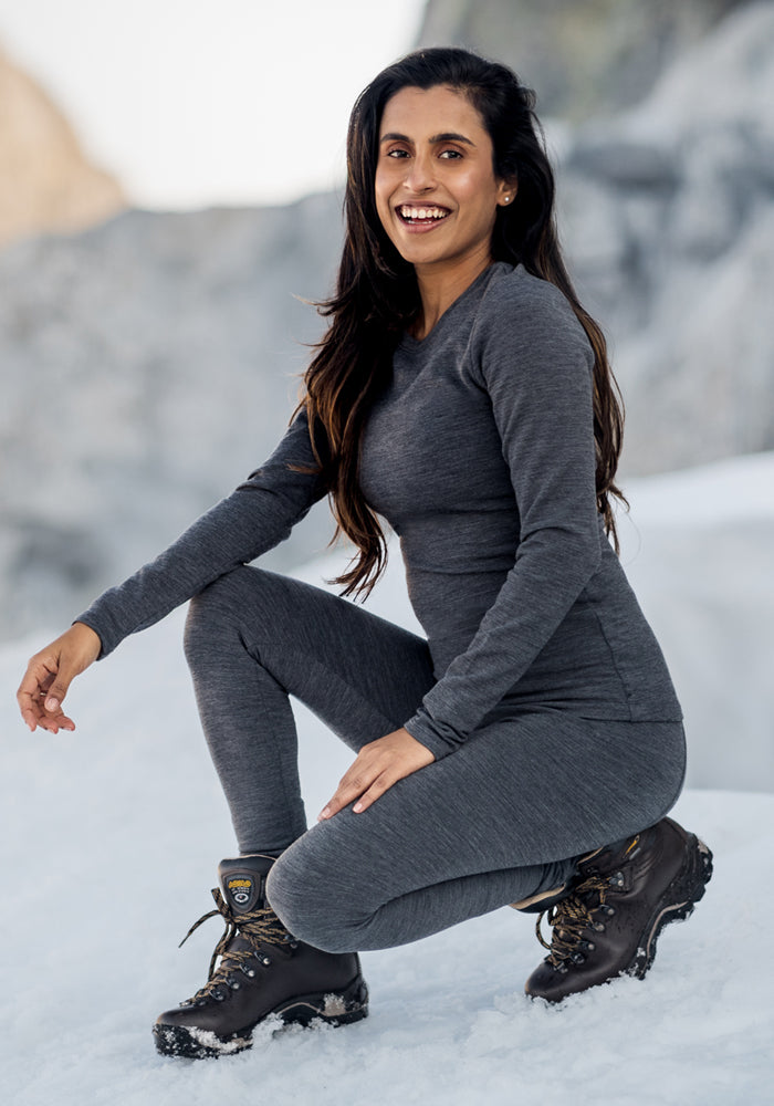 A woman in Woolx Stella Petite - Cinder Melange leggings and hiking boots kneels on snowy ground, smiling at the camera with mountains in the background.