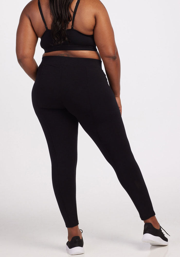 A person is standing with their back to the camera, modeling the Stella Tall - Black leggings by Woolx. These sleek leggings are made from DuraLite fabric and are perfectly paired with a black sports bra and stylish black sneakers featuring white soles, all set against a plain white background.