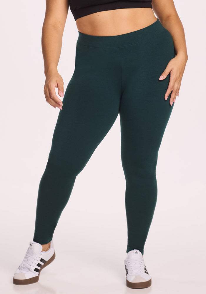Wearing Woolx Stella Tall leggings in Deep Timber (final sale), a person stands against a light background, pairing them with a black crop top and white Adidas sneakers, one hand on their hip and the other by their side.