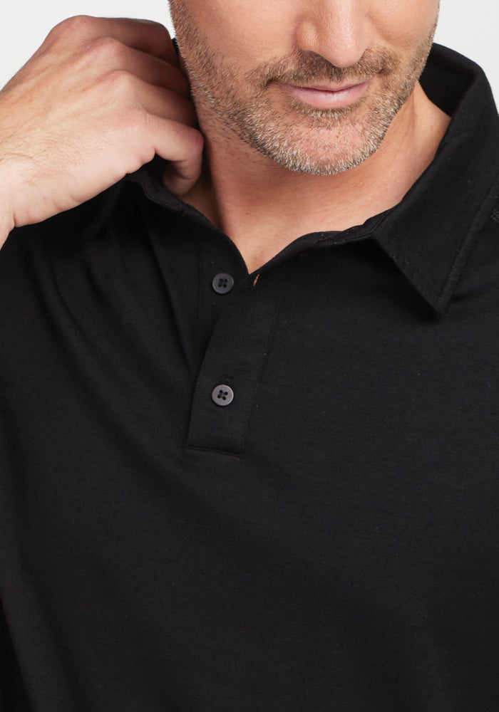 A man with a short beard is adjusting the collar of his Woolx Summit Polo in black, designed as high-performance athletic gear. With two buttons undone, he gazes slightly downward. The image emphasizes the moisture management feature of the shirt while subtly focusing on his upper torso and face without revealing all facial features.
