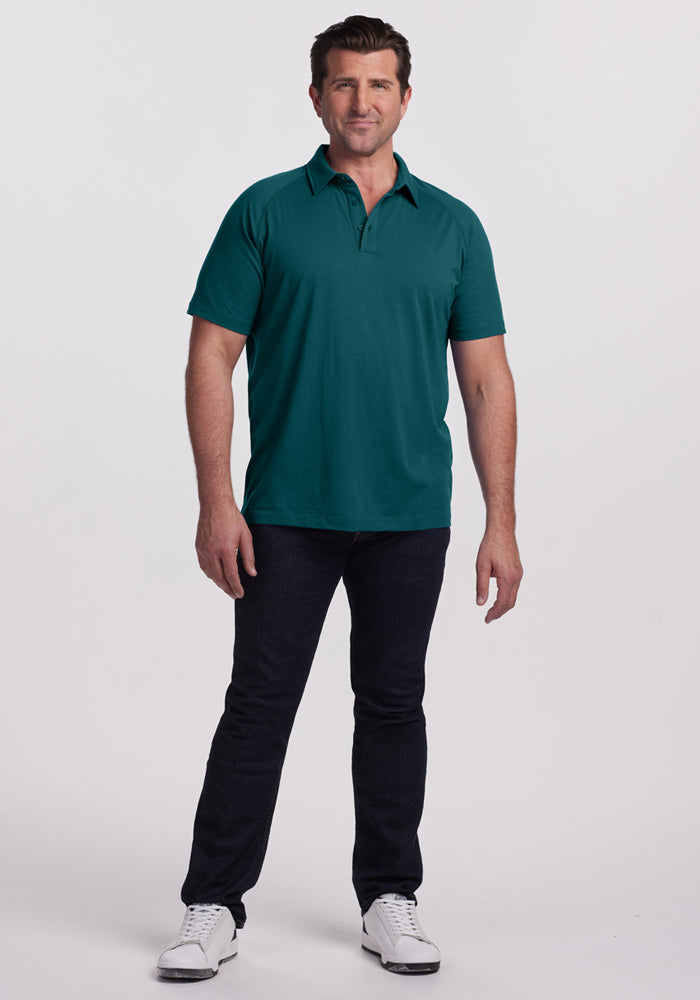 Man smiling, wearing Woolx Summit Polo • Final Sale - Riverbank Green, dark jeans, and white sneakers on white background.