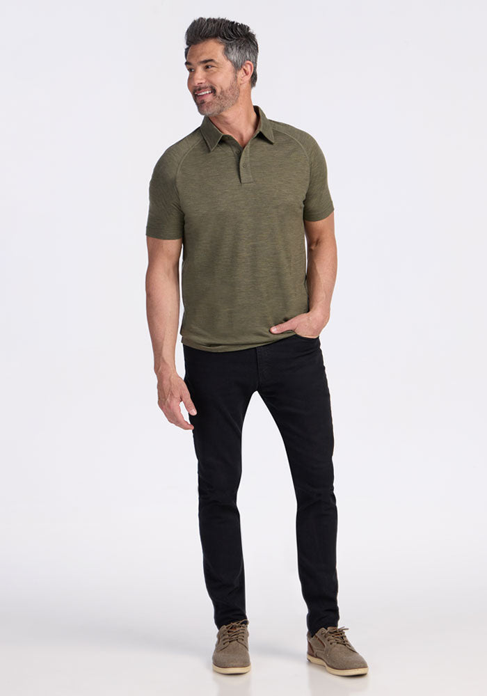 Man wearing the Woolx Summit Polo in Sage Olive Melange, black jeans, and tan shoes, smiling against a white background.