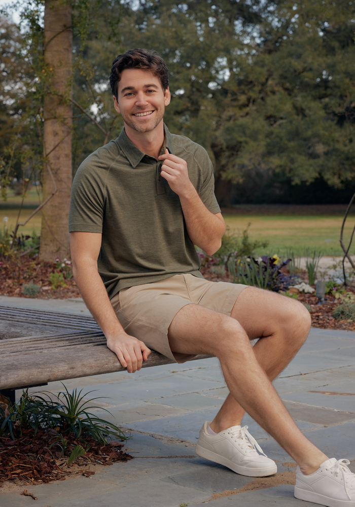 A smiling man wears the Woolx Summit Polo • Final Sale - Sage Olive Melange, sitting outdoors on a park bench.