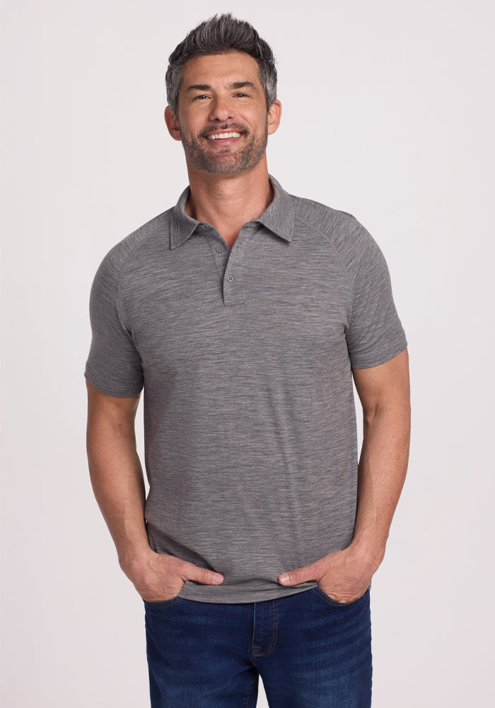 A man with short hair and a beard wears the Woolx Summit Polo in Graphite Heather with jeans, hands in pockets, smiling at the camera against a plain white background, highlighting the shirt's natural moisture management qualities. | Matthew is 6’, wearing a size L