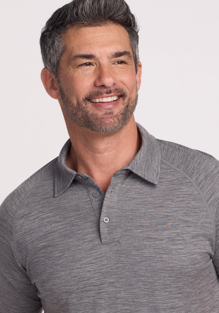 A man with short, graying hair and a beard smiles in a Woolx Summit Polo - Graphite Heather, reflecting the classic warmth and comfort of Merino wool. The plain white background highlights his relaxed demeanor.