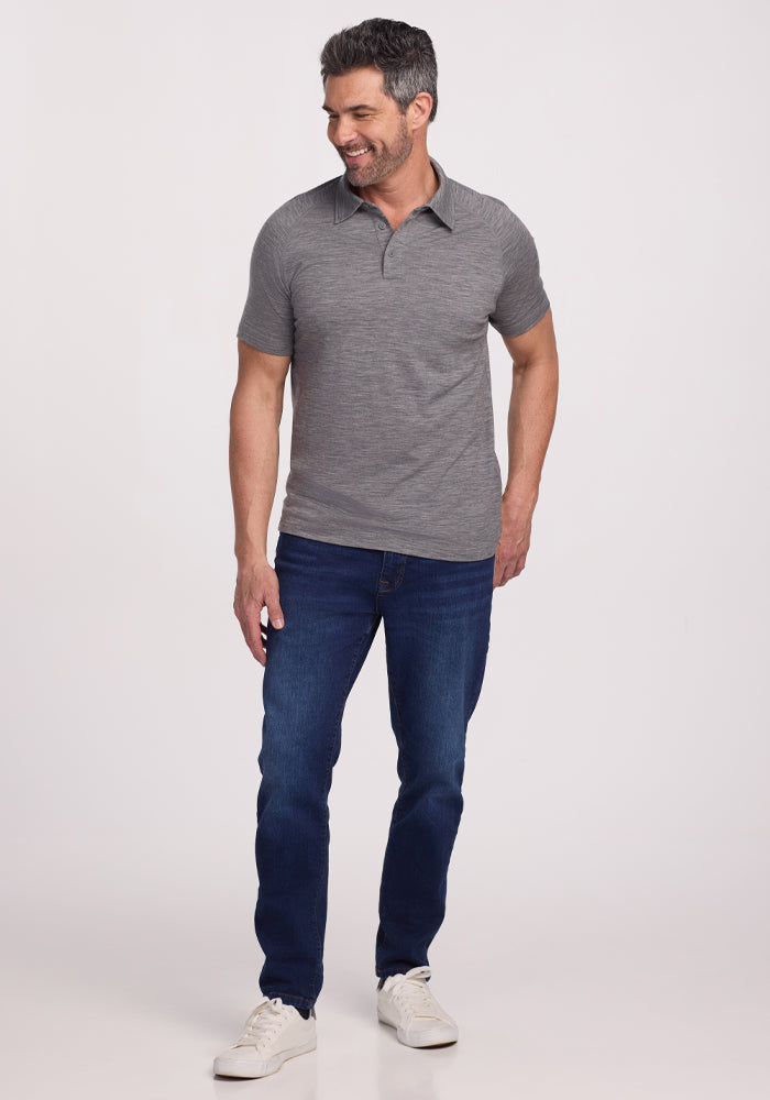 A man stands against a plain background wearing the Woolx Summit Polo in Graphite Heather, dark blue jeans, and white sneakers. Made from Merino wool, the polo offers excellent moisture management. He smiles while looking slightly to the side, with one hand in his pocket.