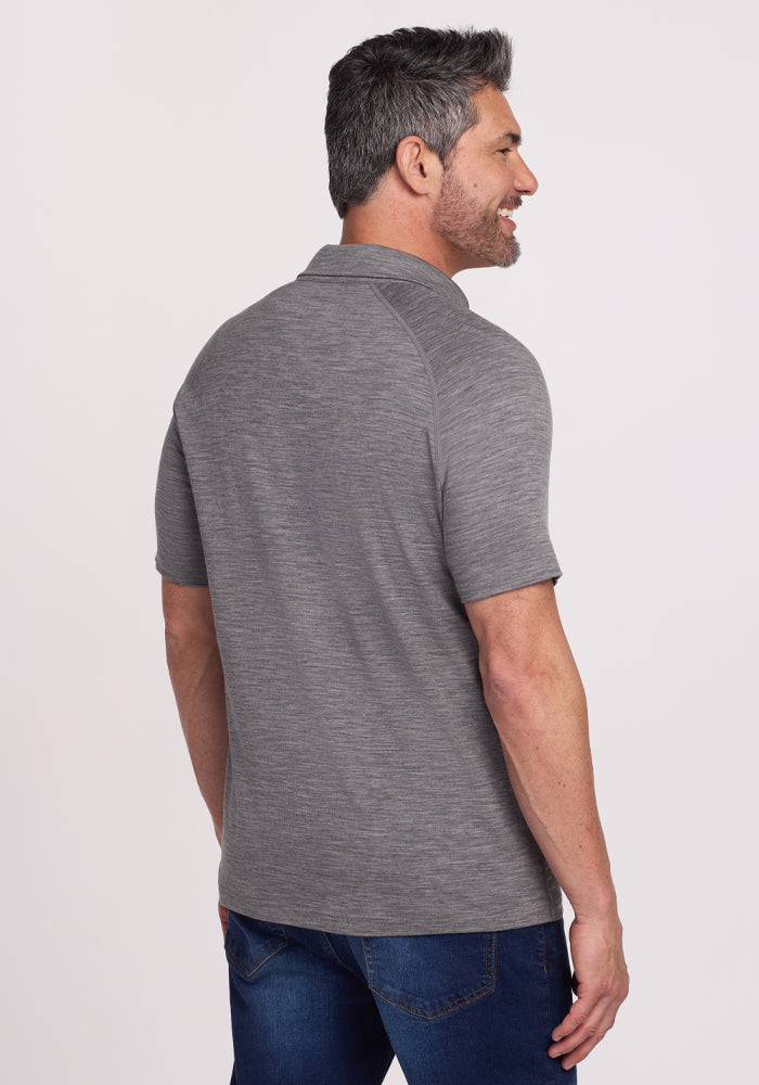 A gray-haired man with a beard smiles while standing sideways, wearing the Woolx Summit Polo in Graphite Heather, known for its moisture management, paired with blue jeans against a plain white background.