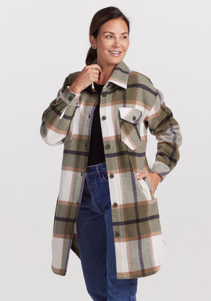 Dressed in the Sutton Shacket - Forest Plaid by Woolx, a person stands confidently with one hand on their hip and the other lightly touching their chin. The shacket features shades of green, brown, and white, offering a breathable plaid design. Complementing this look is a black top and blue jeans, all tied together with a subtle smile.