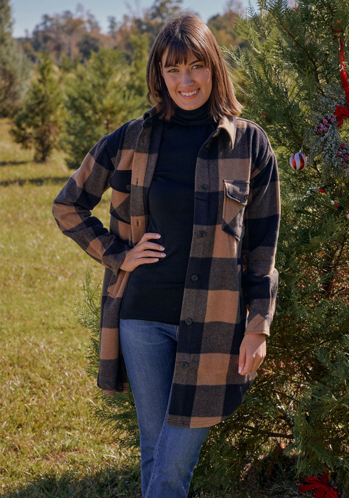 A woman in a black turtleneck, blue jeans, and the Woolx Sutton Shacket • Final Sale - Camel Checkered stands outdoors by a decorated evergreen tree, smiling on a sunny day.
