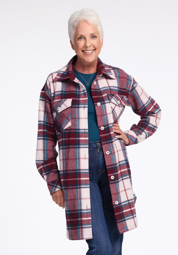 An older woman with short white hair smiles, wearing the Woolx Sutton Shacket in Frozen Cranberry Plaid over a teal top and blue jeans. She poses against a plain white background with one hand on her hip.