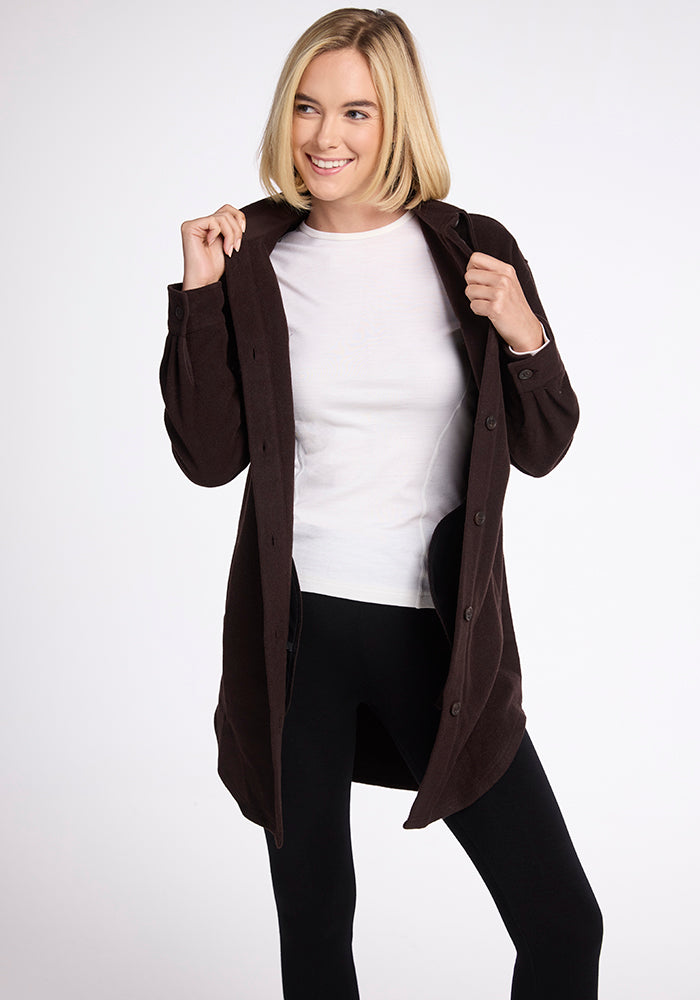 A person wearing a white shirt, black leggings, and the Woolx Sutton Shacket in French Roast smiles while holding the coat's lapels. The background is plain white.