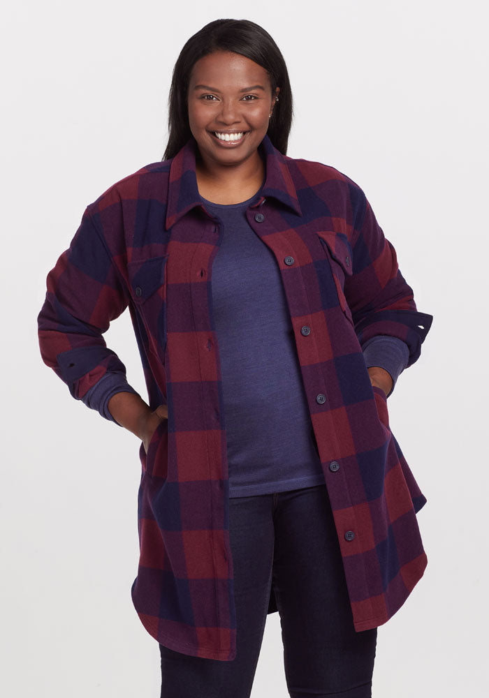 A person smiles in the Woolx Sutton Shacket, featuring a cranberry navy checkered pattern over a dark blue top, hands in pockets. The background is plain light color.