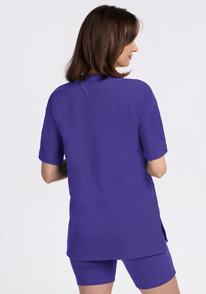 A woman with brown hair wears the Woolx Vera Ribbed Tunic in Berry Indigo, standing with her back to the camera.