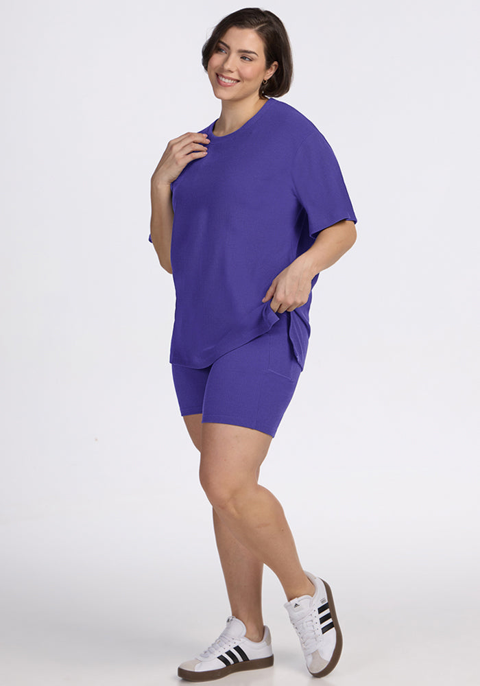 Woman wearing the Woolx Vera Ribbed Tunic in Berry Indigo with shorts and white sneakers, standing and smiling.