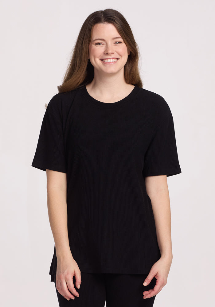 A woman with long brown hair smiles while wearing the Woolx Vera Ribbed Tunic in black and black pants, standing against a plain white background.