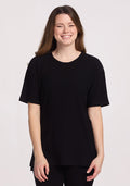 A woman with long brown hair smiles while wearing the Woolx Vera Ribbed Tunic in black and black pants, standing against a plain white background.