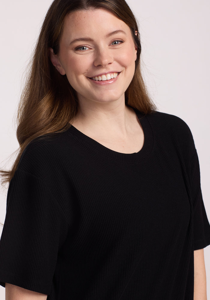 A woman with long brown hair smiles at the camera, wearing the Woolx Vera Ribbed Tunic in black. She stands against a plain, light background.