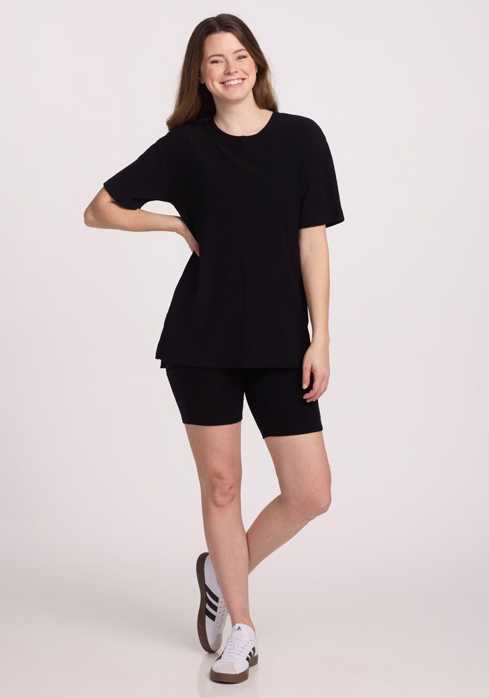A woman with long brown hair stands against a plain light background, smiling in the Woolx Vera Ribbed Tunic - Black, paired with black bike shorts and white sneakers featuring black stripes.