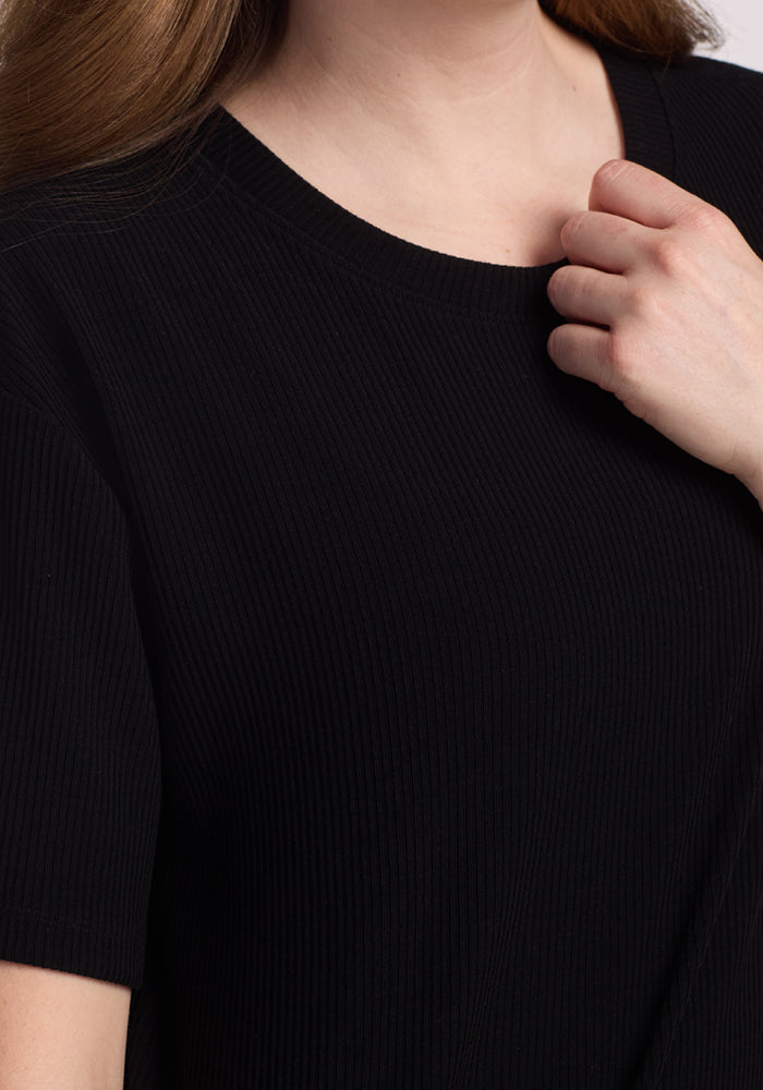 A person wearing the Woolx Vera Ribbed Tunic in black touches the neckline with their right hand, showing only the lower face, neck, and upper torso.